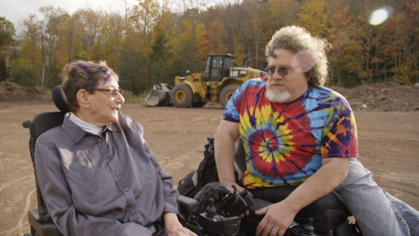 Jim LeBrecht and Denise Jackobson (both in power chairs) on empty campgrounds with bulldozer behind.