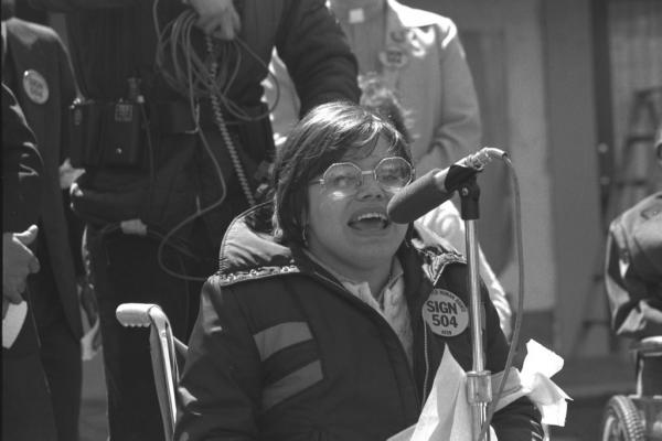 Judy Heumann speaking into a microphone during a rally, wearing a "Sign 504" button.