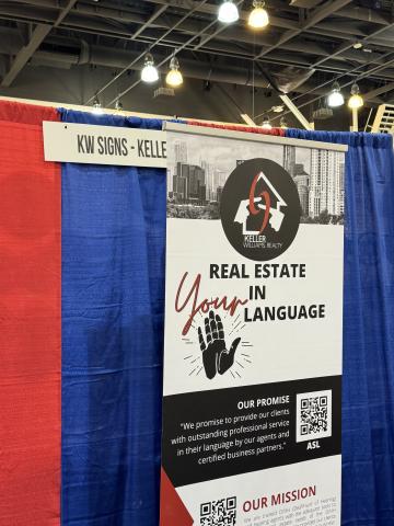 A banner advertising Keller Williams Realty in front of red and blue curtains, banner says “Real estate in your language” with a graphic of an open-faced hand depicting the sign for “your.” The banner has a QR code that connects individuals to the company’s website.