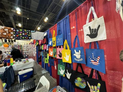 A Deaf-owned business at the Expo selling different sign language-based gifts. Various colored tote bags with the sign for “love” on them hang all across the red and blue curtains.