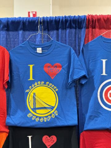 A blue T-shirt with the Warriors logo, a yellow circle with a blue Golden Gate bridge outline, and sign language signs that spell out “Golden State Warriors.” The T-shirt is hung on blue and red curtains. The sign for “love” is shown in a red heart to complete the message of the shirt: “I love the Golden State Warriors.”